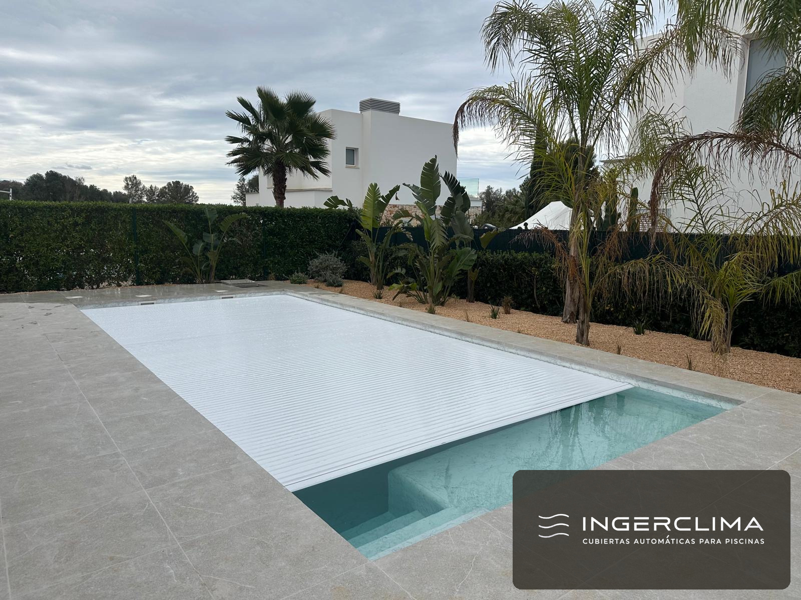 Automatic white slatted cover closed over a villa pool surrounded by palm trees
