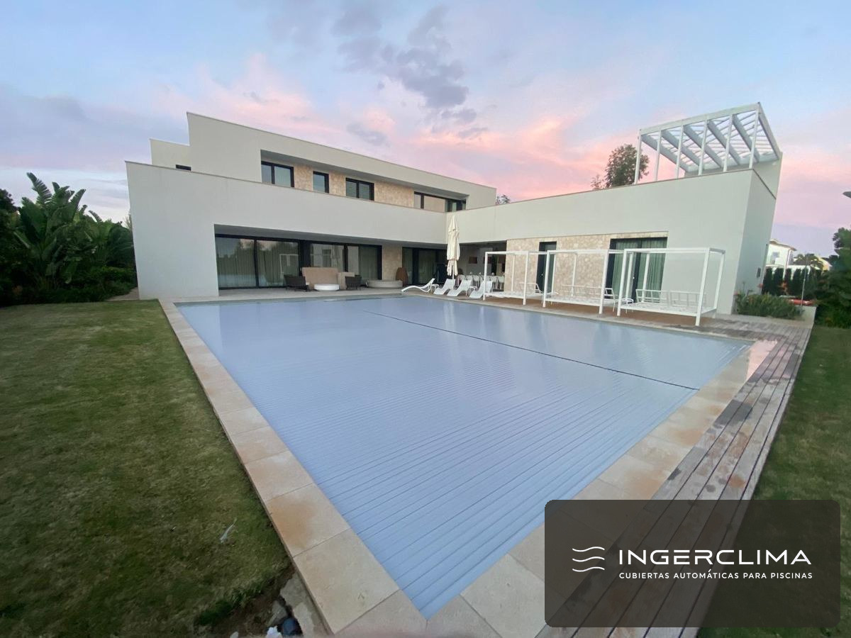 Cubierta automática doble sobre una piscina de gran tamaño, con la vivienda al fondo al atardecer