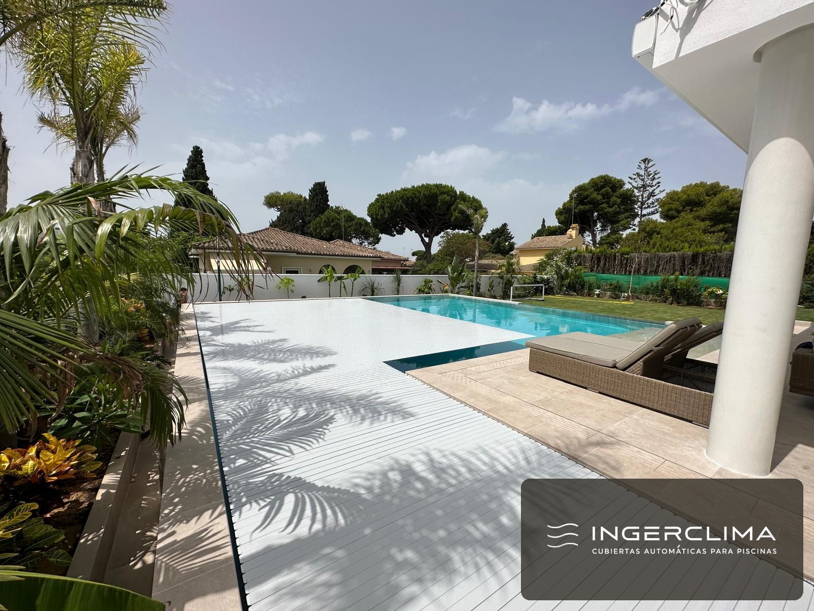 Cubierta automática de lamas blancas cerrada sobre la piscina de una villa, con palmeras y seto alrededor