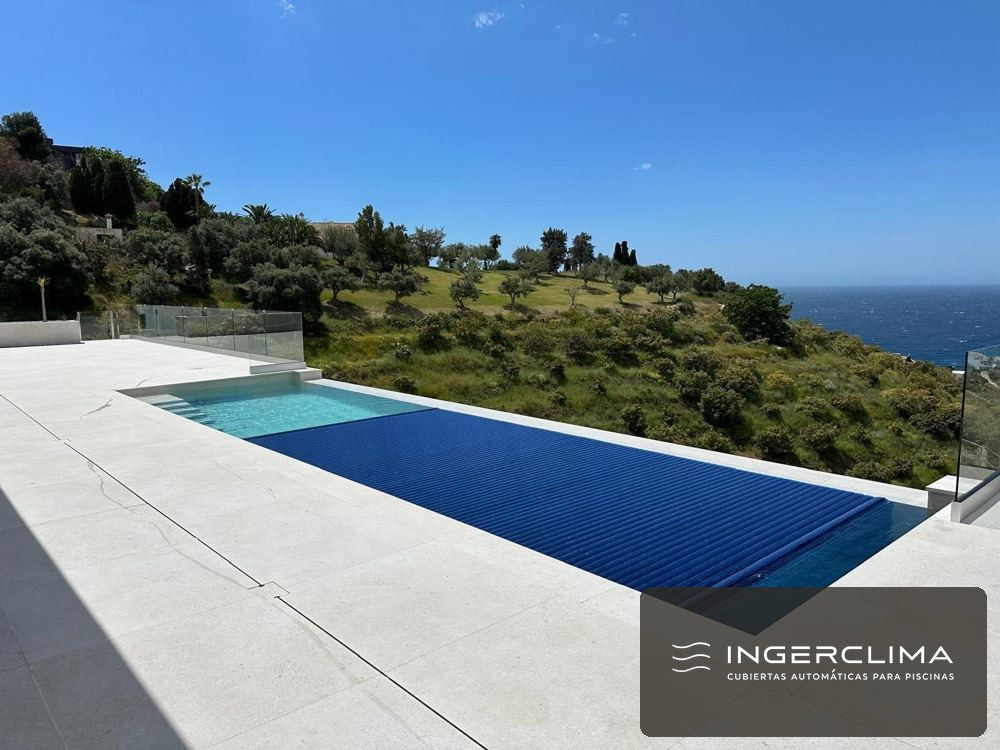 Piscina con la cubierta de lamas a medio recoger al atardecer, en una parcela con jardín
